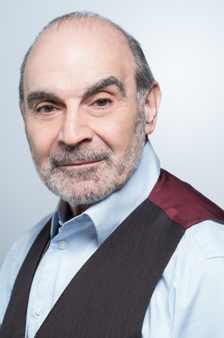 David Suchet A middle-aged man with a bald head and grey beard, wearing a light blue shirt and dark waistcoat with a maroon accent, smiles gently at the camera against a plain light background.
