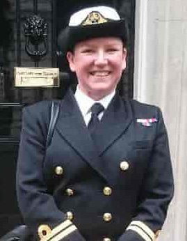 Karen Bassett A smiling person in a dark navy military uniform and hat stands in front of a door with a brass nameplate.