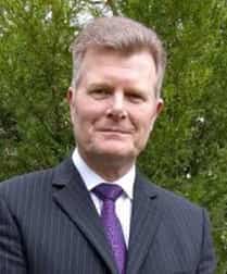 Philip McCormack A man in a dark pinstripe suit and purple tie stands outdoors in front of green foliage, looking at the camera and smiling slightly.