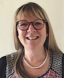Carey Rivers A woman with straight, light brown hair and fringe, wearing round glasses, a pearl necklace, and a patterned top, smiling at the camera against a plain light background.