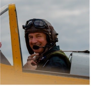 Christopher Huchstep A smiling pilot wearing a vintage leather helmet and goggles is seated in the cockpit of a yellow aeroplane, looking towards the camera against a cloudy sky background.