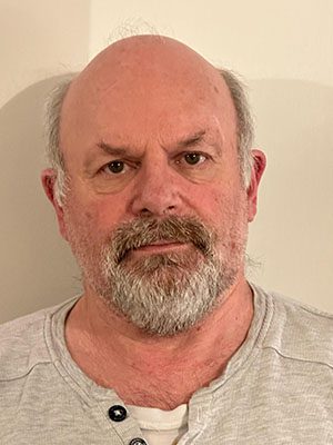 David Cordey A middle-aged man with a bald head, grey beard, and moustache is wearing a light-coloured, buttoned shirt and standing against a plain, light-coloured wall.