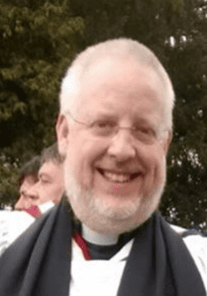 James Pitkin A smiling man with grey hair, glasses, and a beard is wearing clergy attire with a black scarf. Trees and another person are visible in the blurred background.