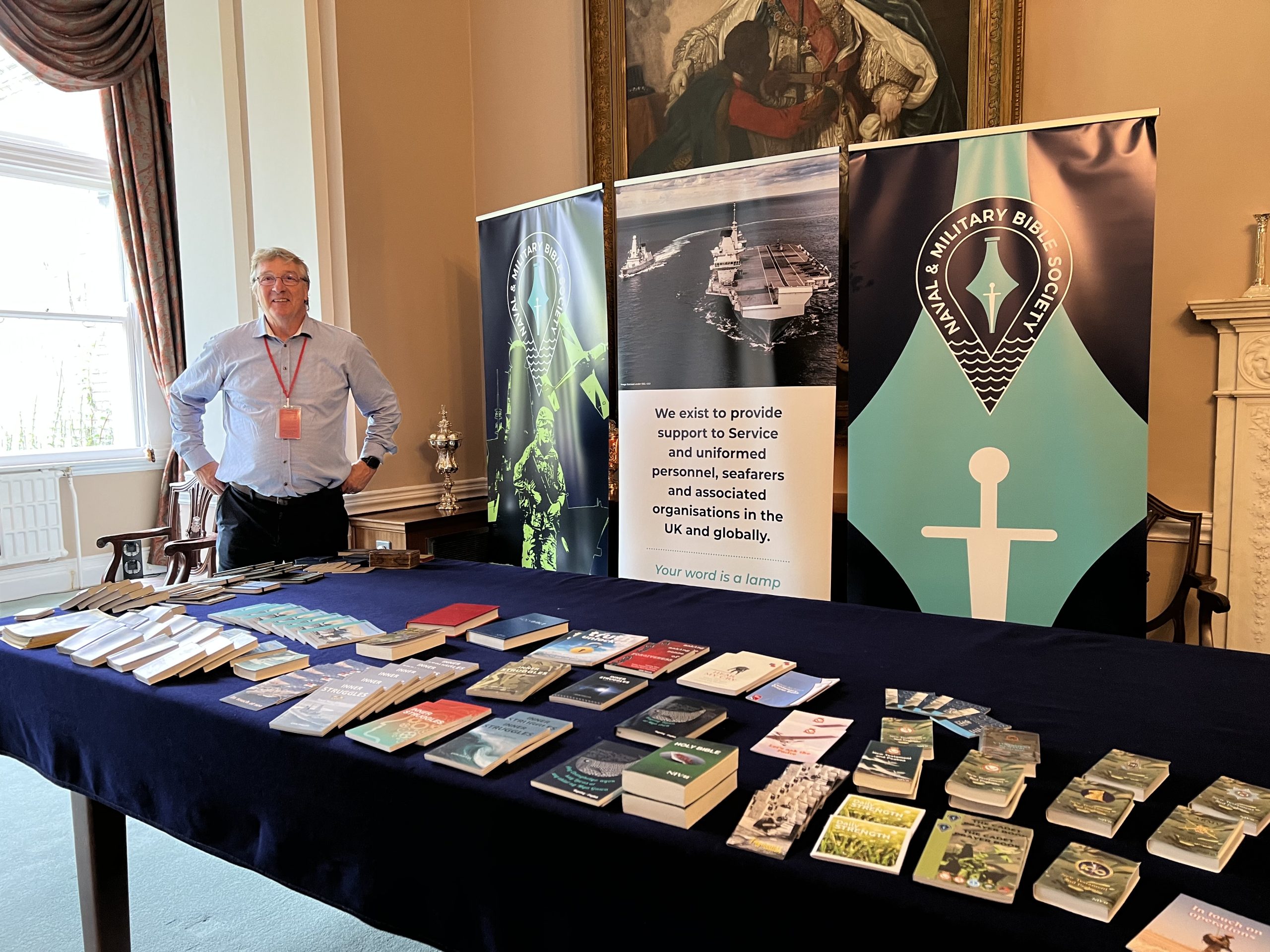 RN Day A man stands smiling behind a table covered with books and pamphlets. Behind him are banners for the Naval Military Bible Society and a sign offering support to service personnel. The setting appears to be a formal room.