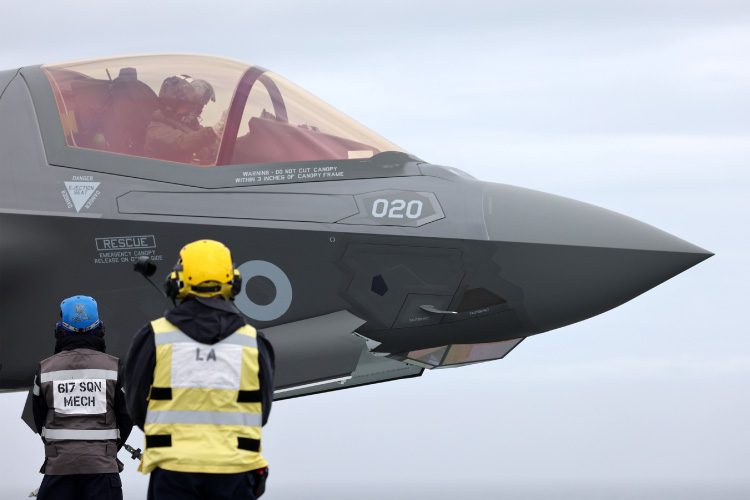 Two ground crew members in helmets and vests stand facing a grey military jet, with a pilot visible inside the cockpit against a cloudy sky. The jet has 020 and a roundel insignia on its nose.