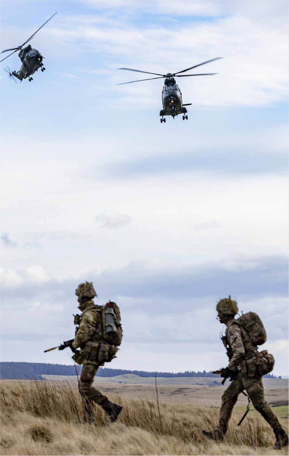 Three soldiers in camouflage gear walk through tall grass as two military helicopters fly overhead in a cloudy sky, with open fields and low hills in the background.