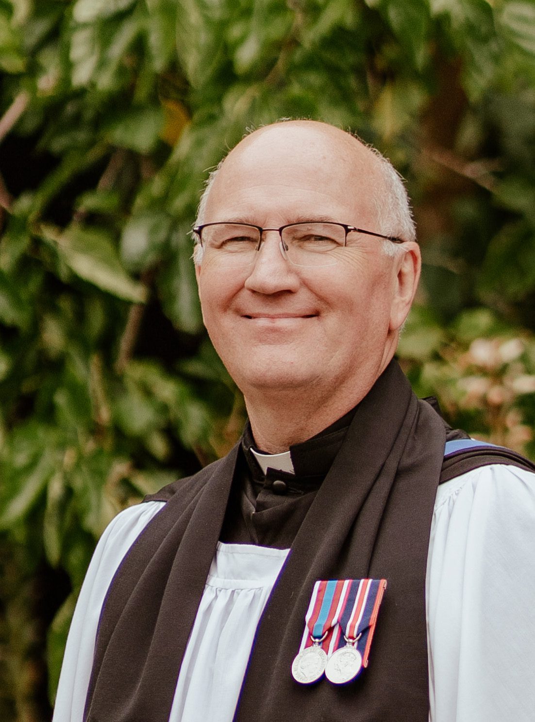 A smiling man with glasses, wearing clerical robes and two medals, stands outdoors in front of leafy greenery. A smiling man with glasses, wearing clerical robes and two medals, stands outdoors in front of leafy greenery.